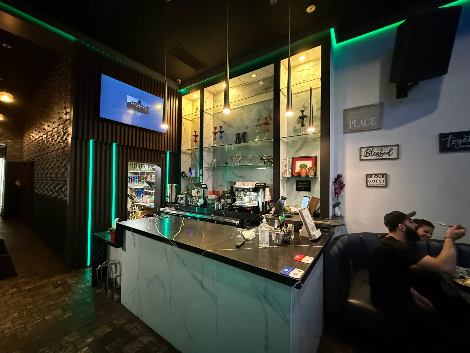 Maestro Lounge bar with a black marble counter, espresso machine, glass shelves of colorful hookah stems, green LED lighting, a wall-mounted screen, and guests seated in blue booths